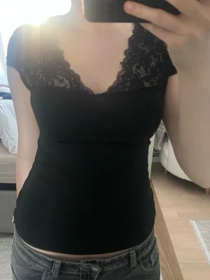 Svart topp med spetsdetaljer - Svart figurnära topp med v-ringning och fina spetsdetaljer vid halsringningen och axlarna. Kortärmad modell som passar perfekt till jeans eller kjol. Snygg och enkel att styla till olika looks.