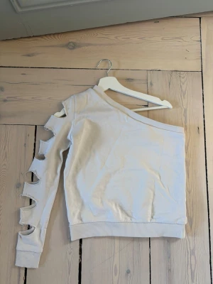 Vit oneshoulder topp från AFJ x NAKD - Säljer en vit oneshoulder topp från AEJ med unik design – ena ärmen är lång och har flera stora utskurna hål längs hela ärmen. Ribbad mudd vid ärmslut och nederkant. Perfekt för dig som vill sticka ut med din stil!