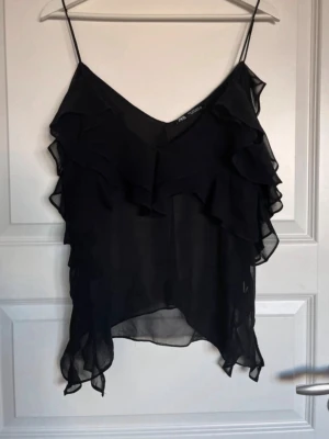 Svart volanglinne från Zara - Svart linne från Zara i tunn, transparent chiffong med volangdetaljer framtill och baktill. Smala axelband och asymmetrisk nederkant ger en luftig och trendig look. Perfekt för lager-på-lager eller som statement-piece.