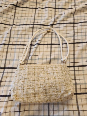 Beige tweed handväska med vit axelrem - Trendig beige handväska i tweed med vit axelrem i repstil och guldfärgade detaljer. Väskan har en mjuk, rektangulär form och stängs med dragkedja. Perfekt accessoar för dig som gillar en chic och stilren look. Den är helt oanvänd och har en fungerande dragkedja. 