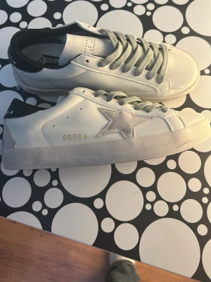 Vita GGDB sneakers med stjärndetalj - Snygga vita sneakers från Golden Goose med ikonisk stjärna på sidan och GGDB-logga i guld. Skorna har grå skosnören, svart hälparti och en slitstark gummisula med fiskbensmönster. Perfekta för dig som gillar streetstyle och vill sticka ut.