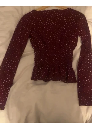 Prickig blus ginatricot - Snygg mörklila blus med vita små prickar och smockad midja som ger en figurnära passform. Blusen har långa ärmar och en lätt volang nertill. Perfekt för dig som gillar trendiga och feminina detaljer.