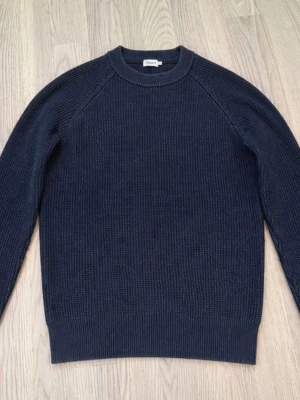 Mörkblå stickad tröja från Filippa K - XS MEN PASSAR MER S. En stilren mörkblå stickad tröja från Filippa K med rund halsringning och ribbade muddar vid ärmslut och nederkant. Perfekt för lager-på-lager och enkel att matcha med jeans eller chinos.