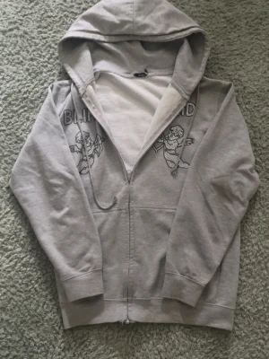 Grå zip hoodie ifrån bling lucid - Grå hoodie med dragkedja från bling lucid . Framsidan har två tryckta änglar och texten 'Bling lucid' i stenar. Mjuk insida, huva och ribbade muddar. Perfekt för en chill och stilren look.