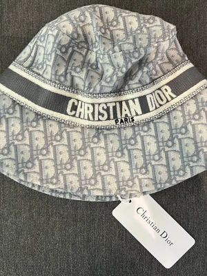 Grå bucket hat från Christian Dior - Snygg bucket hat från Christian Dior i grått med ikoniskt Dior-mönster och bred rand med logotypen broderad runt hatten. Tillverkad i ett mjukt tyg som ger en avslappnad och trendig look. Perfekt accessoar för att lyfta din outfit.