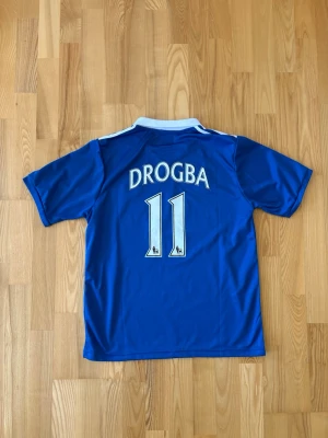 Chelsea Drogba matchtröja Adidas - Blå Chelsea fotbollströja med vitt tryck, Drogba 11 på ryggen. Adidas-logga, Champions League-märke och Samsung som sponsor framtill. Klassisk krage och korta ärmar. Perfekt för fans av fotboll och Chelsea. Står att det är storlek XL men är mer som Medium.