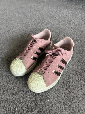 Adidas Superstar rosa sneakers - Säljer ett par Sdidas Superstar Metallic 3 Stripes – Pink Tintsneakers i rosa mocka med klassiska svarta ränder i metallic och vit tåhätta. Dem är använda men fortfarande väldigt fina!  Pris kan diskuteras! 