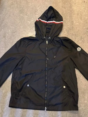 Svart vindjacka från Moncler - Snygg svart vindjacka från Moncler med huva och dragsko. Jackan har dragkedja framtill, två fickor med tryckknappar och Moncler-logga på ärmen. Detaljer i rött och vitt på huvan ger en sportig look. Tillverkad i lätt, vindtätt material.
