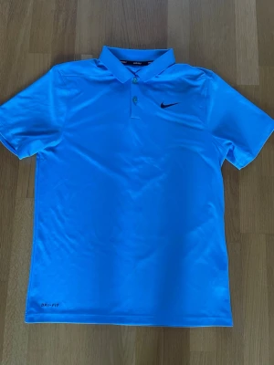 Blå pikétröja från Nike Golf - Snygg ljusblå pikétröja från Nike Golf med klassisk krage och knappar framtill. Tröjan har svart Nike-logga på bröstet och är tillverkad i ett lätt och svalt polyestermaterial med Dri-Fit-teknologi. Nypris 500-600kr