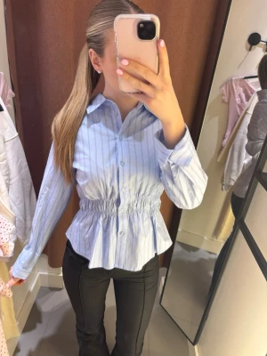 Blå randig skjorta med markerad midja - Snygg blå skjorta med tunna vita ränder och markerad midja med resår. Skjortan har klassisk krage, knappar framtill och lång ärm. Perfekt för en trendig och stilren look. Endast använd 1 gång