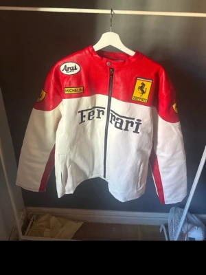 Ferrari racinginspirerad skinnjacka - Racinginspirerad jacka i skinn med Ferrari-logga och patchar från Aral och Michelin. Jackan är röd och vit med svarta detaljer, dragkedja framtill och tryck på både fram- och baksida. Perfekt för dig som gillar motorsport och vill sticka ut.