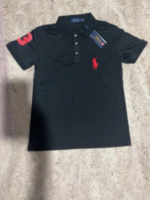 Svart pikétröja från Polo Ralph Lauren - Klassisk svart pikétröja från Polo Ralph Lauren med röd broderad logga på bröstet och stor röd siffra på ärmen. Tröjan har krage, knappar framtill och är gjord i mjuk bomull. Perfekt för en clean och sportig look. Storlek kan passa upp till M