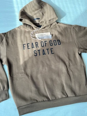Fear of God Essentials hoodie brun - Snygg brun hoodie från Fear of God Essentials med texten 'FEAR OF GOD STATE' tryckt framtill. Klassisk passform med huva och ribbade muddar. Tillverkad i mjukt bomullsmaterial för maximal komfort. Perfekt för en avslappnad och trendig streetwear-look.