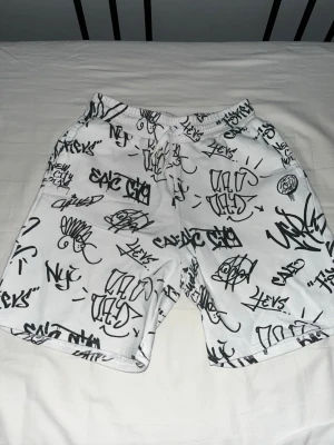 Vita shorts med graffiti-tryck - Säljer ett par vita shorts med svart graffiti-inspirerat tryck över hela plagget. Shortsen har elastisk midja med snörning och avslappnad passform. Perfekta för dig som gillar streetwear och vill sticka ut lite extra. Storlek: 158 EUR