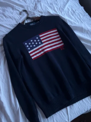 Ralph lauren tröja stickad  - Snygg mörkblå stickad tröja från Ralph Lauren med en stor amerikansk flagga på bröstet och RL-broderi. Tröjan har rund halsringning och ribbade muddar vid ärmslut och nederkant. Perfekt för en cool och avslappnad stil.