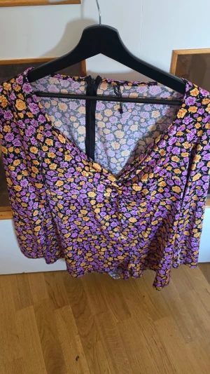 Blommig body med v-ringning - Body med färgglatt blommönster i lila, gult och svart. V-ringad framtill och med långa ärmar. Tunt, lätt material som ger ett luftigt intryck. Perfekt för dig som gillar färg och mönster.