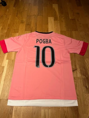 Juventus away kit Pogba #10 - Juventus matchtröja i rosa med Pogba och nummer 10 på ryggen. Tröjan har svarta Adidas-ränder på axlarna, Jeep-logga på bröstet och klubbmärke. Tillverkad i lätt och ventilerande climacool-material. Perfekt för fotbollsträning eller som supporterplagg.