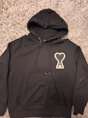 Svart hoodie från Ami Paris - Svart hoodie från Ami Paris med stor vit broderad logga i form av ett hjärta och bokstaven A på bröstet. Perfekt för dig som vill han en fin hoodie. Den är inte använd så mycket.