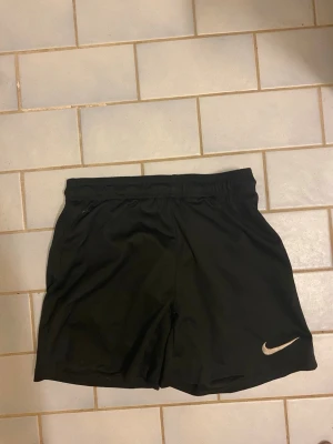 Svarta träningsshorts från Nike - Svarta träningsshorts från Nike med elastisk midja och klassisk vit Nike-logga på vänster ben. Tillverkade i lätt och snabbtorkande polyester, perfekta för sport och aktivitet. Enkel och stilren design som funkar till det mesta.