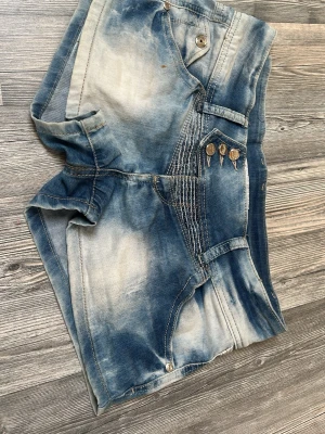 Blå tie-dye jeansshorts med detaljer - Säljer ett par blå tie-dye jeansshorts från Siviglia med coola sömdetaljer och tre dekorativa knappar fram. Shortsen har fickor både fram och bak samt bälteshällor. Materialet är denim och färgen är en mix av ljusblått och vitt.