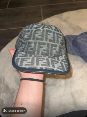 Fendi keps med FF-mönster - Snygg keps från Fendi med ikoniskt FF-mönster i grå och svarta toner. Kepsen har böjd skärm med mörk kant och justerbar rem baktill. Tillverkad i textilmaterial och har en klassisk streetwear-vibe. Perfekt för dig som gillar exklusiva accessoarer.