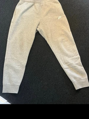 Gråa mjukisbyxor från Nike - Snygga grå mjukisbyxor från Nike med klassisk logga på vänster lår. Byxorna har resår i midjan och vid bensluten för en skön passform. Perfekta för chill eller träning. Tillverkade i mjukt material som känns bekvämt hela dagen.