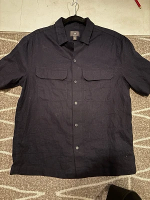 Svart kortärmad skjorta från H&M - Stilren svart skjorta från H&M med regular fit. Skjortan har korta ärmar, två bröstfickor med lock och knäppning framtill. Tillverkad i ett svalt linnematerial som passar perfekt för varmare dagar. Enkel och snygg att matcha med olika outfits.