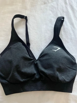 Svart sport-bh från Gymshark - Säljer en svart sport-bh från Gymshark med justerbara axelband och diskret logga på vänster sida. Bh:n är tillverkad i en blandning av nylon och elastan, vilket ger en mjuk och stretchig känsla. Perfekt för träning eller vardag.