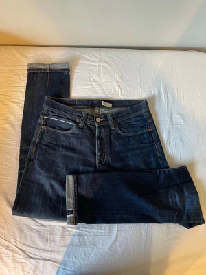 Mörkblå selvedge denim jeans - Säljer ett par mörkblå jeans med raka ben och klassisk femficksmodell. Jeansen har kontrastsömmar i gult och uppvikta benslut för en snygg detalj. Tillverkade i slitstark denim som passar perfekt till din vardagsstil.