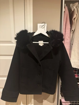 Svart kappa med fuskpäls från Gina Tricot - Stilren svart kappa från Gina Tricot med huva och fuskpälsdetaljer. Kappan har två stora fickor framtill, knäppning med knappar och en rak, något kortare passform. Perfekt för kyliga dagar när du vill vara både varm och snygg.