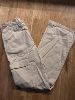 Beige cargobyxor Bershka  - Snygga beige cargobyxor från Bershka med raka ben och flera praktiska fickor på sidorna och bak. Byxorna har bälteshällor och klassisk cargomodell, perfekta för en avslappnad streetstil. Tillverkade i mjukt bomullstyg.