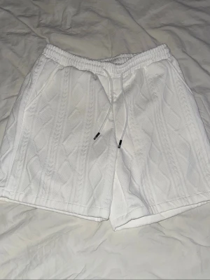 Vita kabelstickade shorts med snörning - Säljer ett par vita kabelstickade shorts med elastisk midja och snörning framtill. Shortsen har en loose passform och är perfekta för en chill och avslappnad stil. Materialet känns mjukt och bekvämt mot huden.