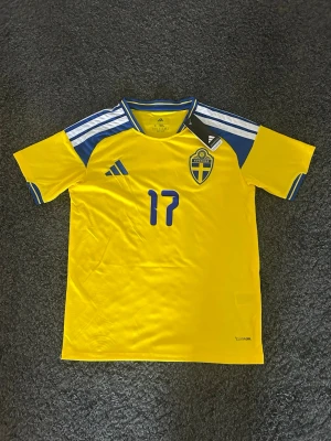 Svensk landslagströja Adidas gul S - Gul svensk landslagströja från Adidas med blå detaljer, nummer 17 på bröstet och klassiskt landslagsemblem. Tröjan har korta ärmar, blå och vita ränder på axlarna och är i slim fit-modell. Perfekt för fotboll eller träning.