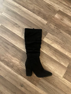 Svarta höga stövlar med klack - Snygga svarta stövlar med hög klack och slouchy skaft. Stövlarna har en dragkedja på insidan och är tillverkade i ett mockaliknande material. Perfekta för att lyfta vilken outfit som helst med sin trendiga och stilrena look.