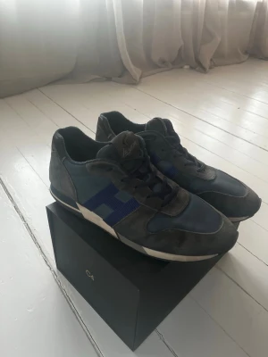 Gråblå sneakers från Hogan - Snygga sneakers från Hogan i grått och blått med mockadetaljer och blått H på sidan. Skorna har klassisk låg profil, snörning och en vit sula som ger en sportig vibe. Perfekta för dig som gillar stilrena och bekväma sneakers. Nypris:- 4000kr