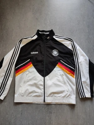 Svart vindjacka från Adidas - Svart vindjacka från Adidas med klassiska vita detaljer och Adidas logga. Jackan har dragkedja framtill och är tillverkad i lätt polyester, perfekt för en sportig och avslappnad stil.