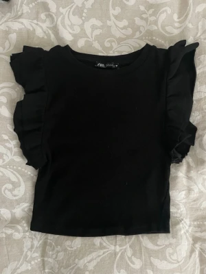 Svart volangtopp från Zara - Snygg svart topp från Zara med volangdetaljer längs axlarna. Modellen är croppad och har en rund halsringning. Storlek M men sitter som XS