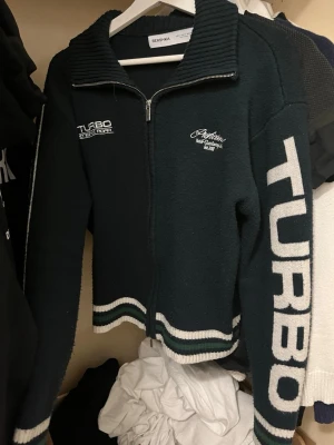 Mörkgrön ziptröja Turbo Bershka - Mörkgrön stickad ziptröja från Bershka med vita detaljer och texten TURBO på ärmen samt Fast Lane Society på ryggen. Tröjan har ribbad krage, dragkedja och sportig collegekänsla. Perfekt för dig som gillar streetwear och unika prints.