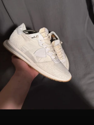 Vita sneakers från Philippe Model - Stilrena vita sneakers från Philippe Model med detaljer i mocka och textil. Skorna har en klassisk låg siluett, vita snören och en beige sula. Perfekta för dig som gillar en clean look och vill ha något som funkar till det mesta.