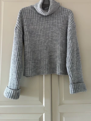 Grå ribbstickad polotröja från NA-KD - Snygg grå ribbstickad polotröja med långa ärmar och uppvikta muddar. Tröjan har en hög polokrage och är gjord i ett mjukt, värmande material. Från NA-KD. Lite ytnopprig, annars är den i fint skick. Storlek S. Lite kort i modellen. 
