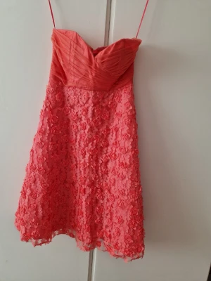 Rosa kort klänning med spetsdetaljer - Superfin rosa kort klänning med tunna axelband och v-ringad överdel. Kjolen är täckt av 3D-blommor i spets och överdelen har snygga veck. Perfekt för sommarens alla tillfällen!