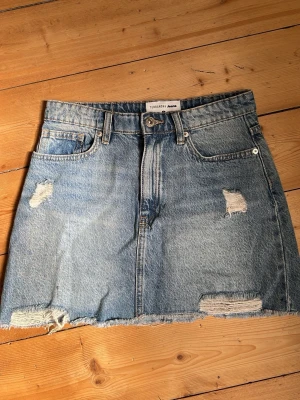 Blå jeanskjol  - Snygg blå jeanskjol med slitna detaljer framtill. Klassisk femficksmodell med bälteshällor. Perfekt för dig som gillar en avslappnad och trendig stil. Knappt använd.