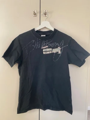 Svart Billabong t-shirt med tryck - Säljer en svart t-shirt från Billabong med coolt tryck och textdetaljer på bröstet. T-shirten har rund hals och korta ärmar, perfekt för en avslappnad streetwear-look. Materialet känns mjukt och skönt, troligen bomull. Passformen är lite mindre än M jag skulle säga att det lutar mer åt S