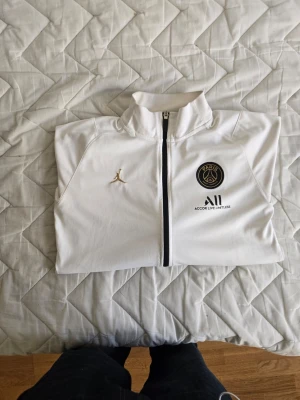 Vit PSG x Jordan träningsjacka - Snygg orginal vit träningsjacka från Jordan och PSG med svart logga på bröstet och guldfärgad Jumpman. Jackan har hel dragkedja, svarta detaljer på ärmarna och två fickor. Perfekt för fotbollsträning eller streetwear.