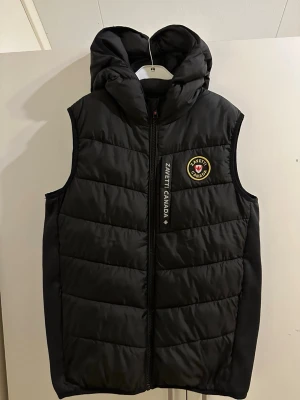 Svart dunväst från Zavetti Canada - Svart dunväst med huva från Zanetti Canada. Västen har ett broderat märke på bröstet, dragkedja framtill och en stor silverfärgad lönnlövsgrafik på ryggen. Snygg och sportig look, perfekt för lager-på-lager-stil.
