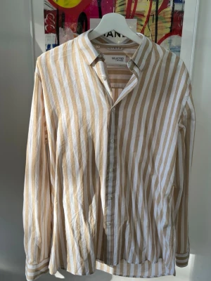 Guldig och vit randig i bomull och linne - Säljer en snygg beige och vit randig skjorta från Selected Homme i luftigt bomull linne. Skjortan har klassisk krage, knappar framtill och långa ärmar. Perfekt för dig som gillar en avslappnad men stilren look.