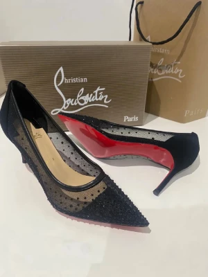 Svarta meshpumps från Louboutin - Snygga svarta pumps från Christian Louboutin med spetsig tå, meshmaterial med prickigt mönster och dekorativa svarta stenar framtill. Klassisk röd sula och låg klack. Perfekta för dig som vill sticka ut med exklusiva detaljer.