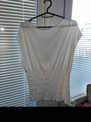 Vit blus med smockad midja - Snygg vit blus med lös passform och smockad midja som ger en cool siluett. Toppen är ärmlös och har en rund halsringning. Perfekt att styla med jeans eller kjol för en fräsch look.