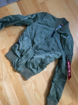 Grön bomberjacka från Alpha Industries - Stilren grön bomberjacka från Alpha Industries med klassisk passform, ribbade muddar och dragkedja framtill. Jackan har en ikonisk röd tag på ärmfickan och två snedställda fickor med tryckknapp. Perfekt för en cool streetstyle-look.