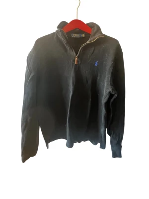 Svart halvzip från Polo Ralph Lauren - Svart långärmad tröja från Polo Ralph Lauren med halv dragkedja och blå broderad logga på bröstet. Hög krage och klassisk passform, perfekt för en stilren look. Materialet känns mjukt och bekvämt.
